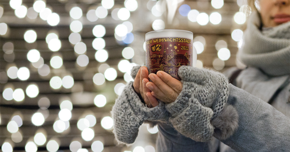 Frau hält mit Glühwein gefüllte, satinierte Tasse in den Händen. Auf der Tasse steht Dortmunder Weihnachtsstadt und es ist ein gezeichneter Druck mit Dortmunder Sehenwürdigkeiten zu sehen.